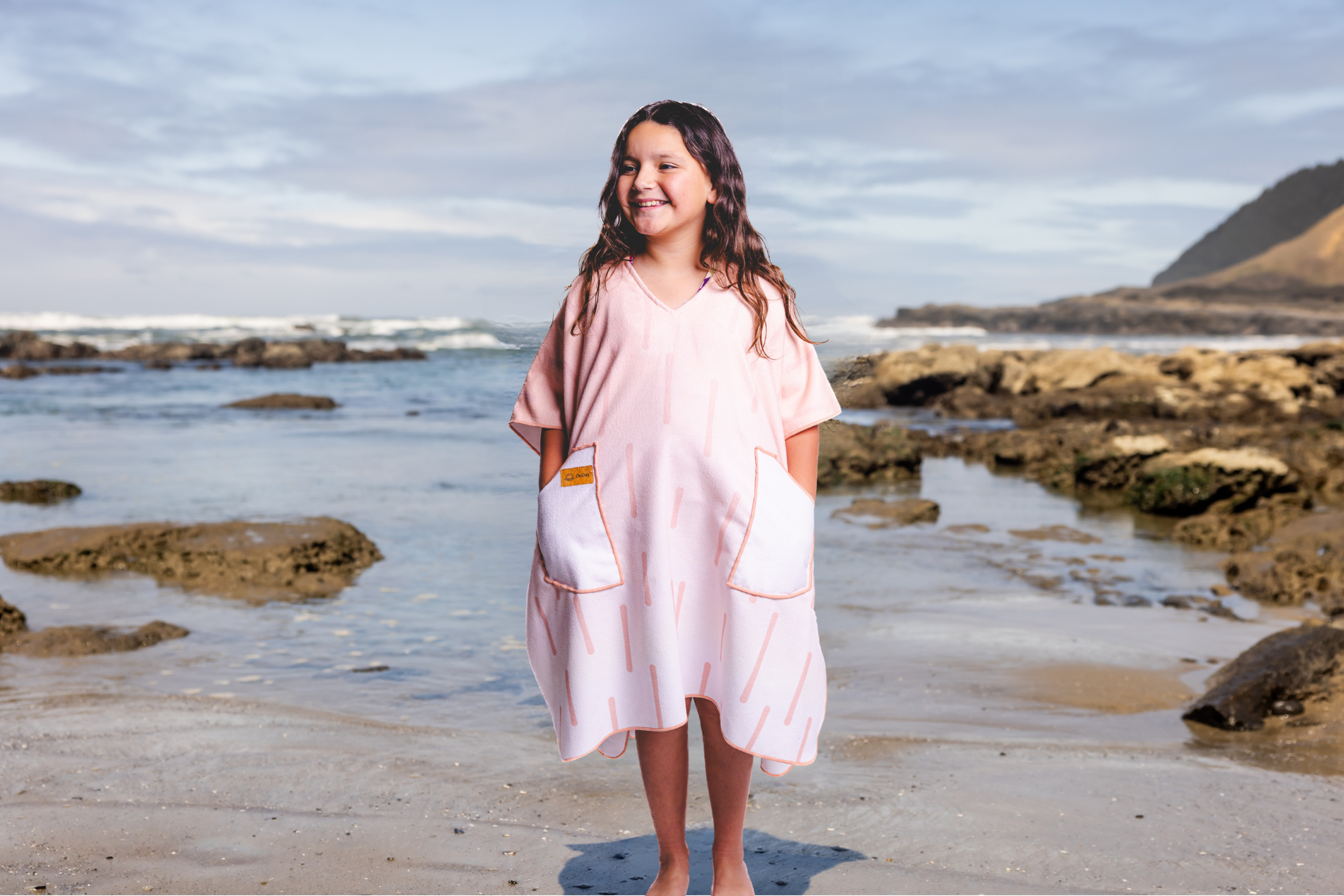 Person on a beach wearing a colorful poncho towel with ocean and rocks in the background