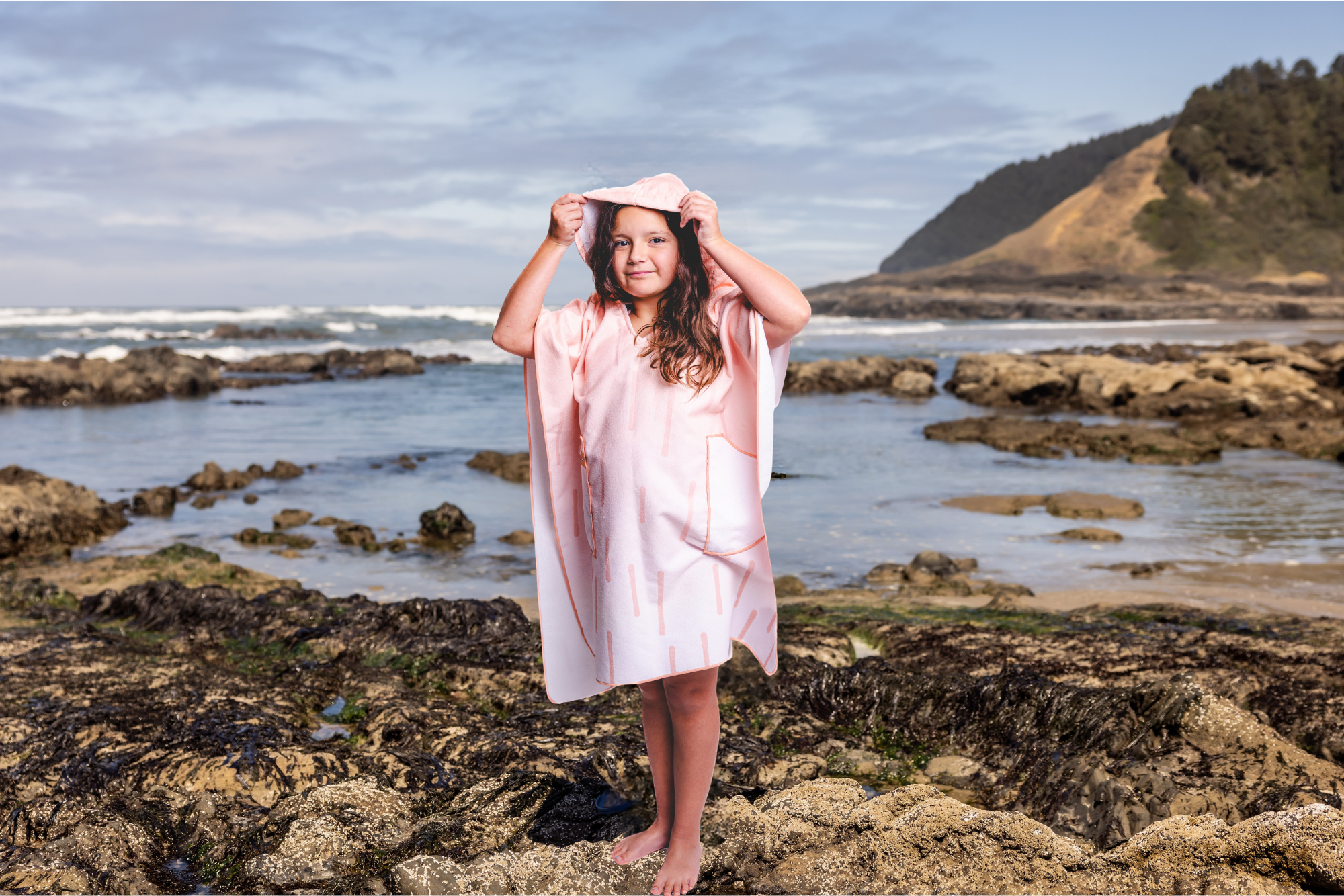 Person on a beach wearing a colorful poncho towel with ocean and rocks in the background