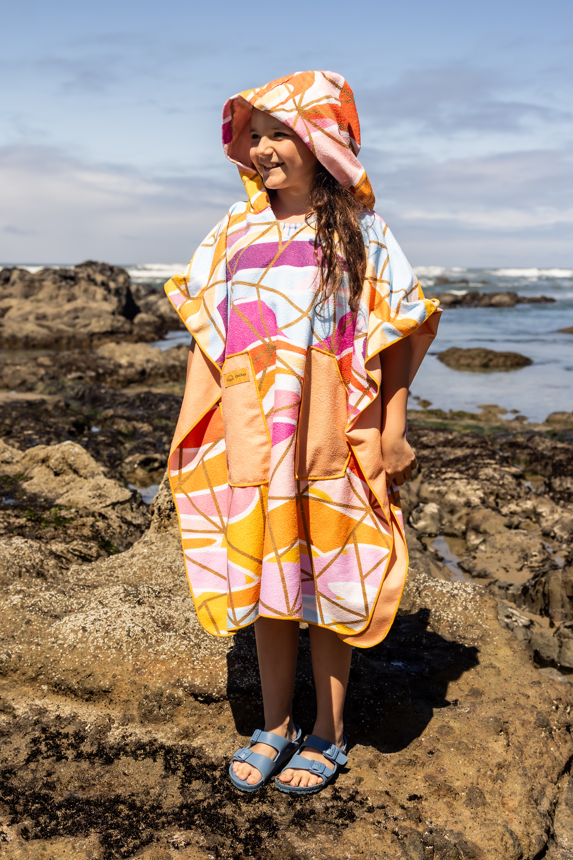 Person on a beach holding a colorful poncho towel with ocean and rocks in the background