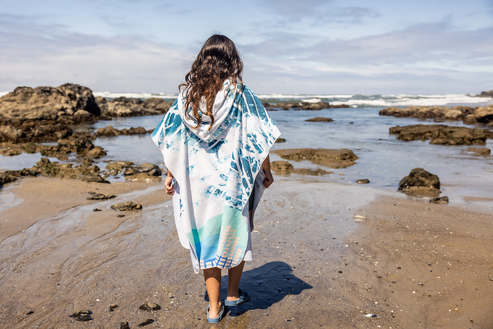 Person on a beach wearing a colorful poncho towel with ocean and rocks in the background