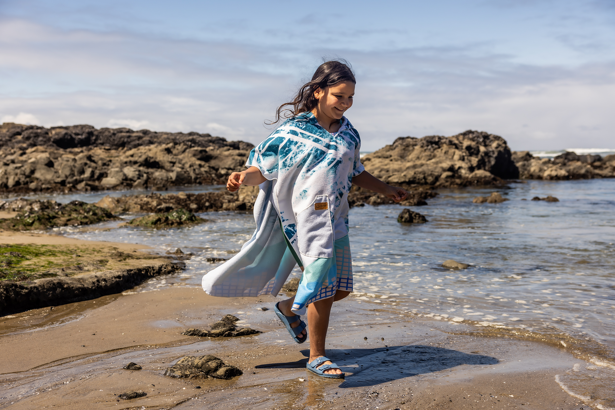 Person on a beach wearing a colorful poncho towel with ocean and rocks in the background