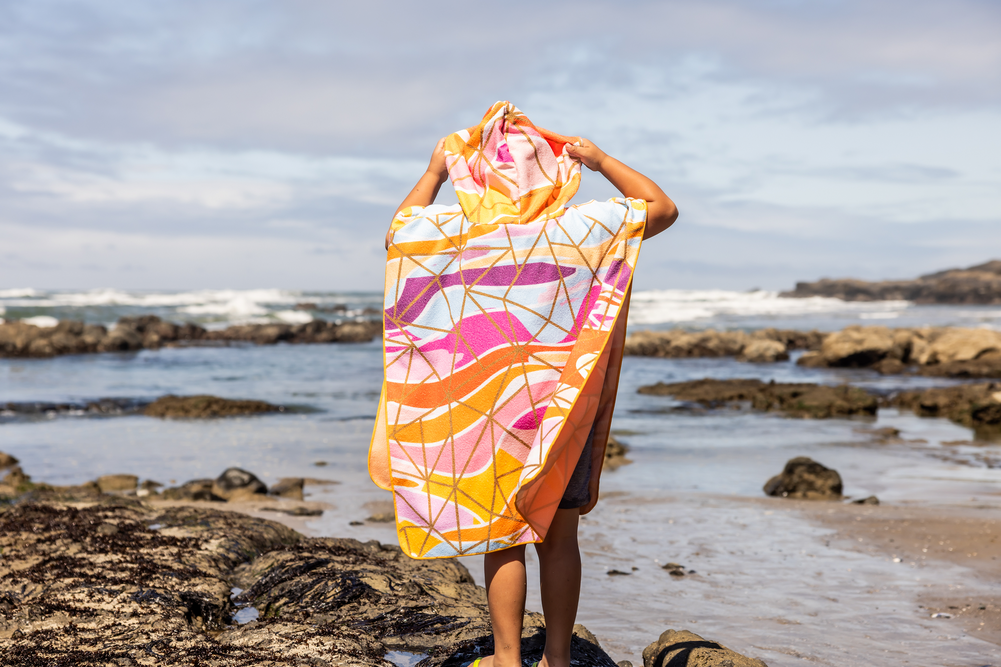 Person on a beach holding a colorful poncho towel with ocean and rocks in the background