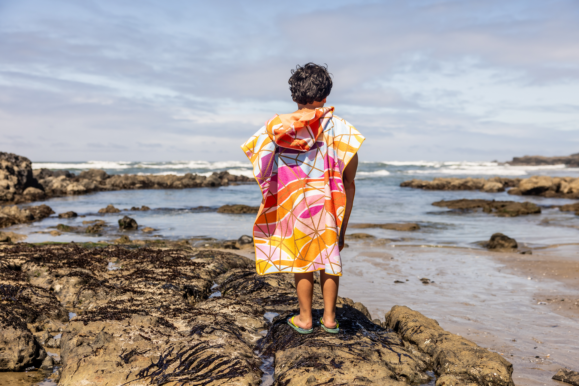 Person on a beach holding a colorful poncho towel with ocean and rocks in the background