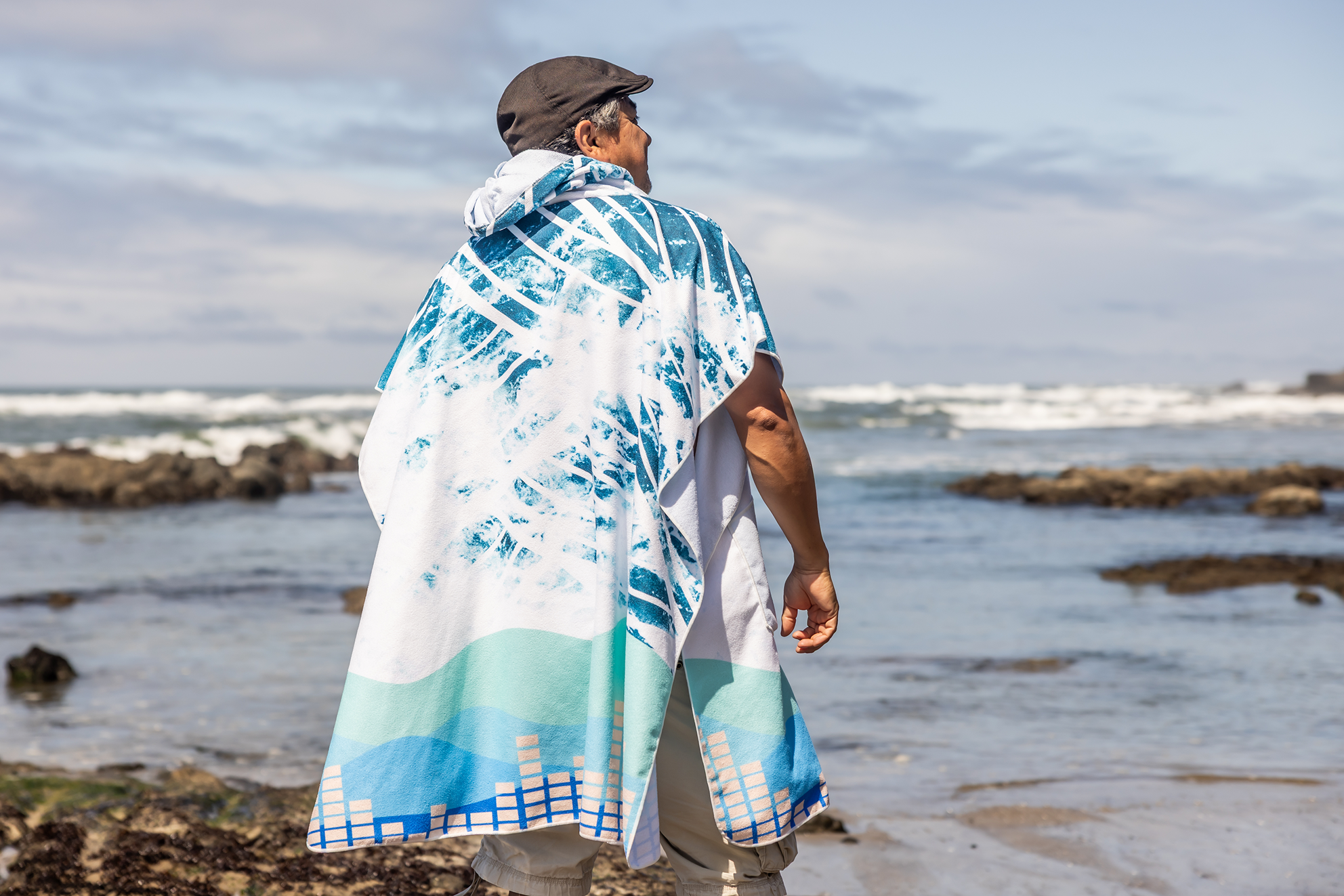 Person on a beach wearing a colorful poncho towel with ocean and rocks in the background