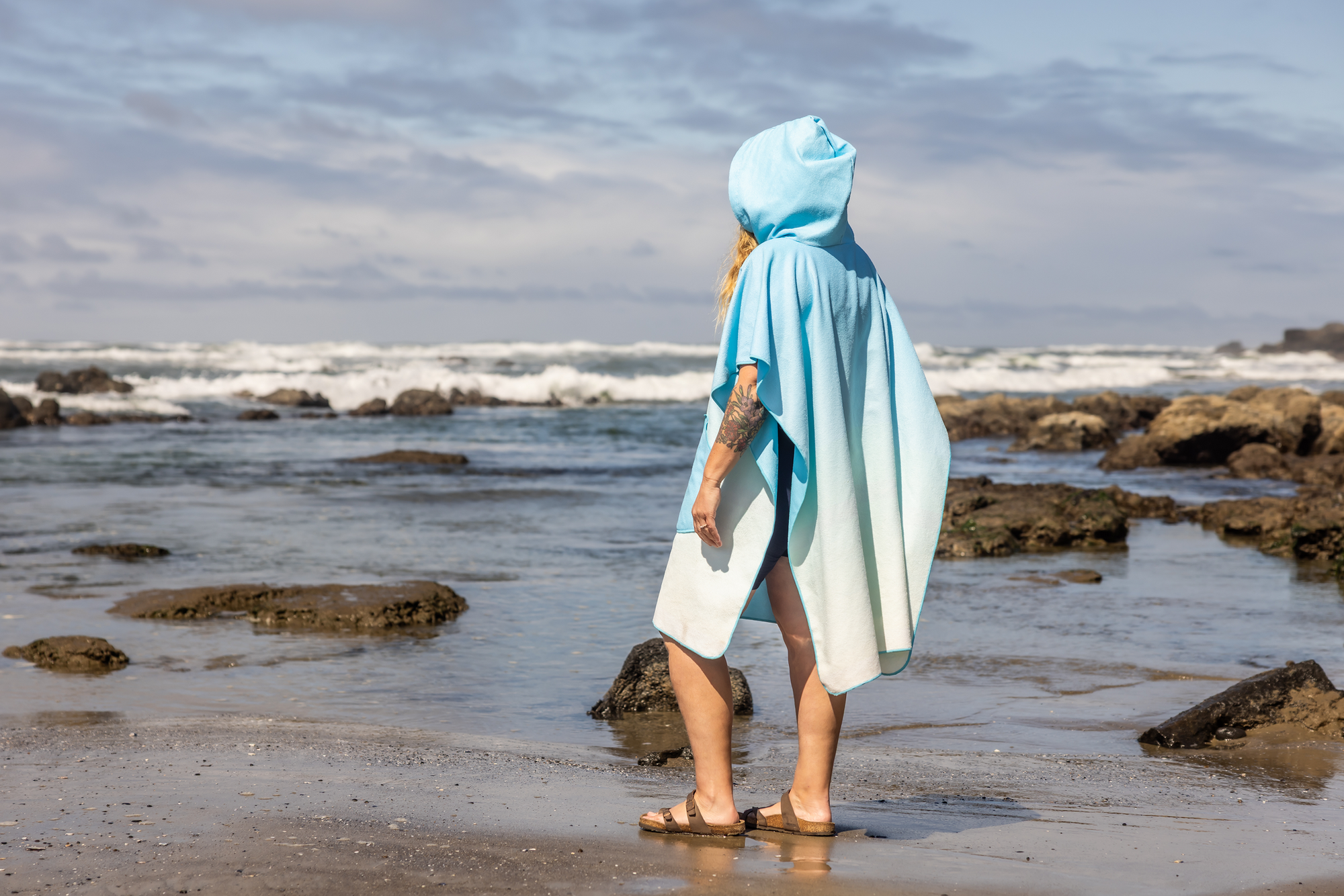 Person on a beach wearing a colorful poncho towel with ocean and rocks in the background