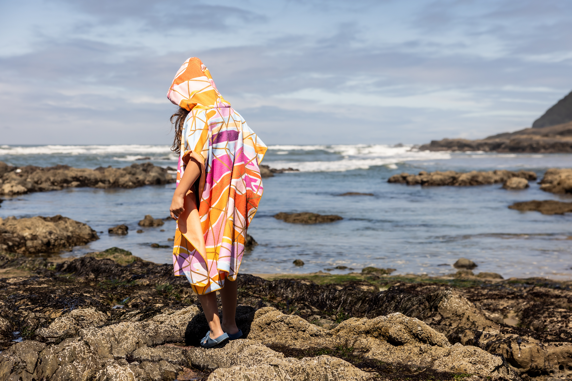 Person on a beach holding a colorful poncho towel with ocean and rocks in the background