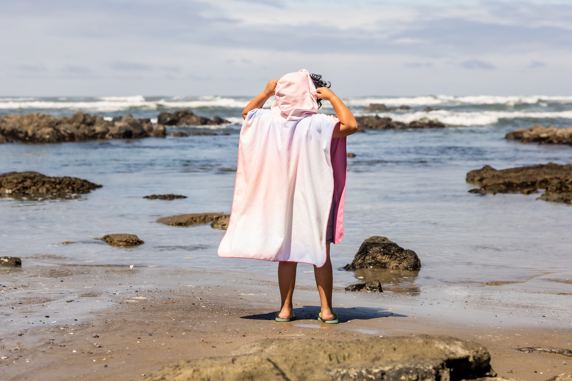 Person on a beach wearing a colorful poncho towel with ocean and rocks in the background