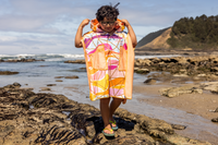 Person on a beach holding a colorful poncho towel with ocean and rocks in the background