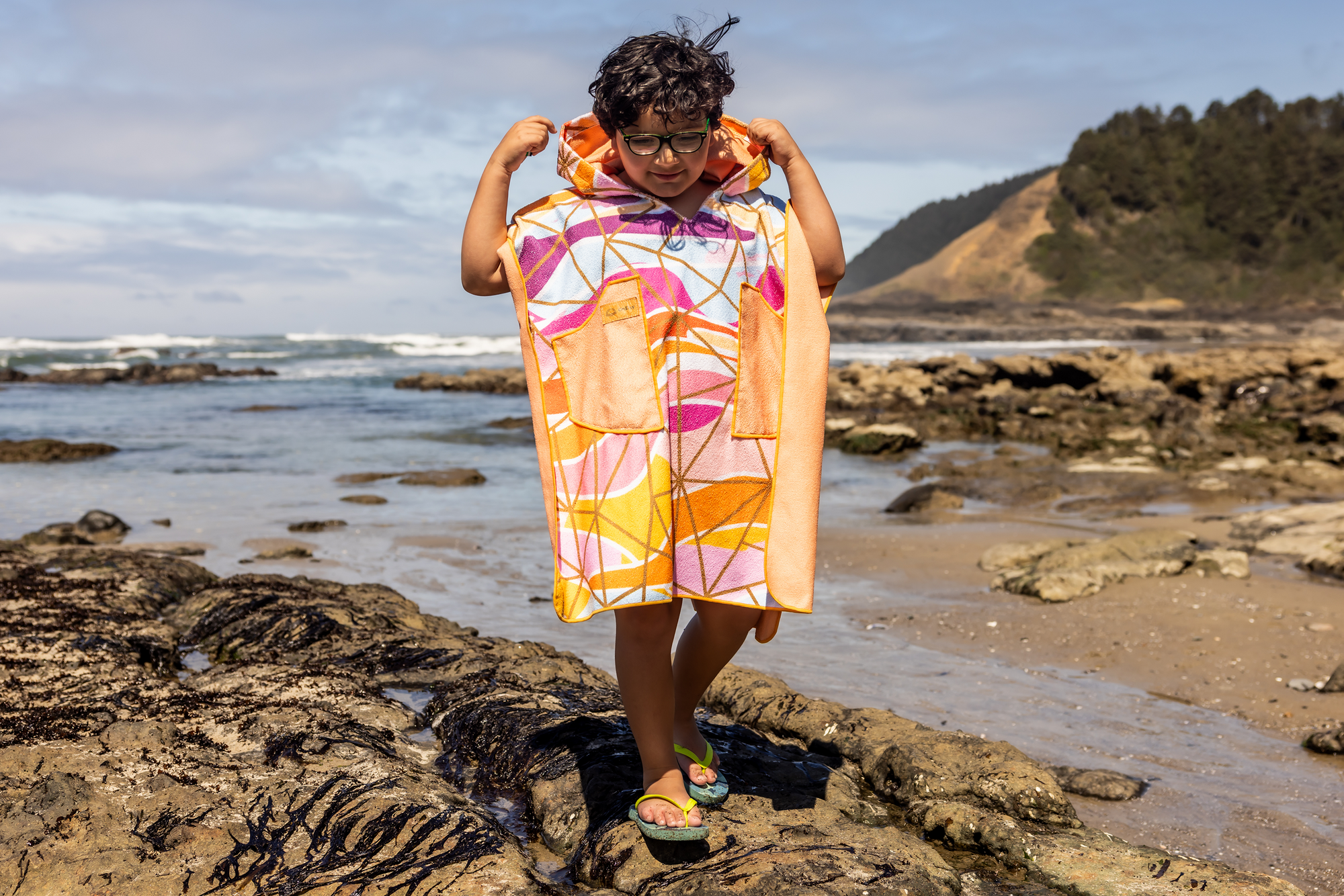Person on a beach holding a colorful poncho towel with ocean and rocks in the background