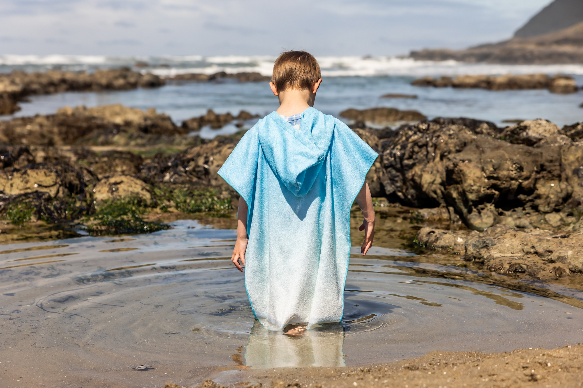 Person on a beach wearing a colorful poncho towel with ocean and rocks in the background