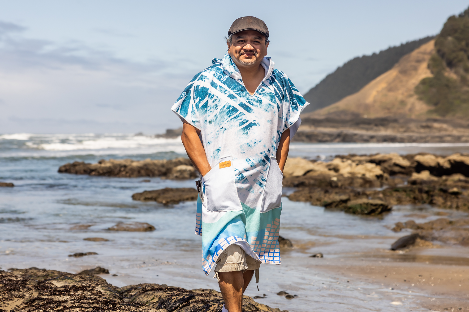 Person on a beach wearing a colorful poncho towel with ocean and rocks in the background